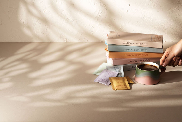Stack of pastel coloured london nootropics coffee boxes on a minimalistic white background with gentle shadows casted from an angle. Next to the stack of boxes is an assortment of London Nootropics adaptogenic coffee blends and a mug full of mushroom coffee.