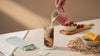 A dining table scene with fruit and a hand pouring milk into an iced coffee while London Nootropics boxes with adaptogenic coffee blends are changing in the background.