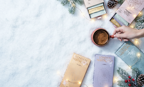 Person holding a mug of hot chocolate with London Nocturne chocolate bars and festive decorations on a snowy background.